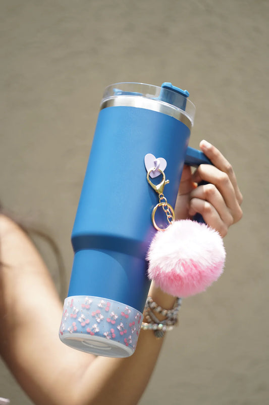 Cherry Blush water bottle boot with a white base and pink cherry-and-bow pattern fitted around the bottom of a blue water bottle, shown outdoors with a fluffy pink charm.