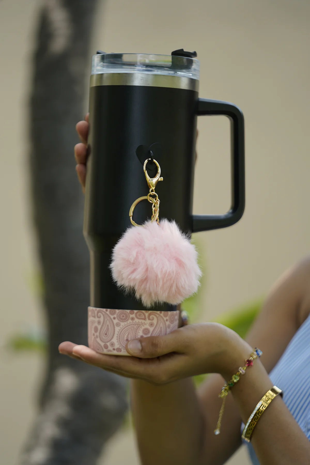 Blush paisley water bottle boot fitted around the base of a black handled water bottle, with a fluffy pink charm, shown outdoors.