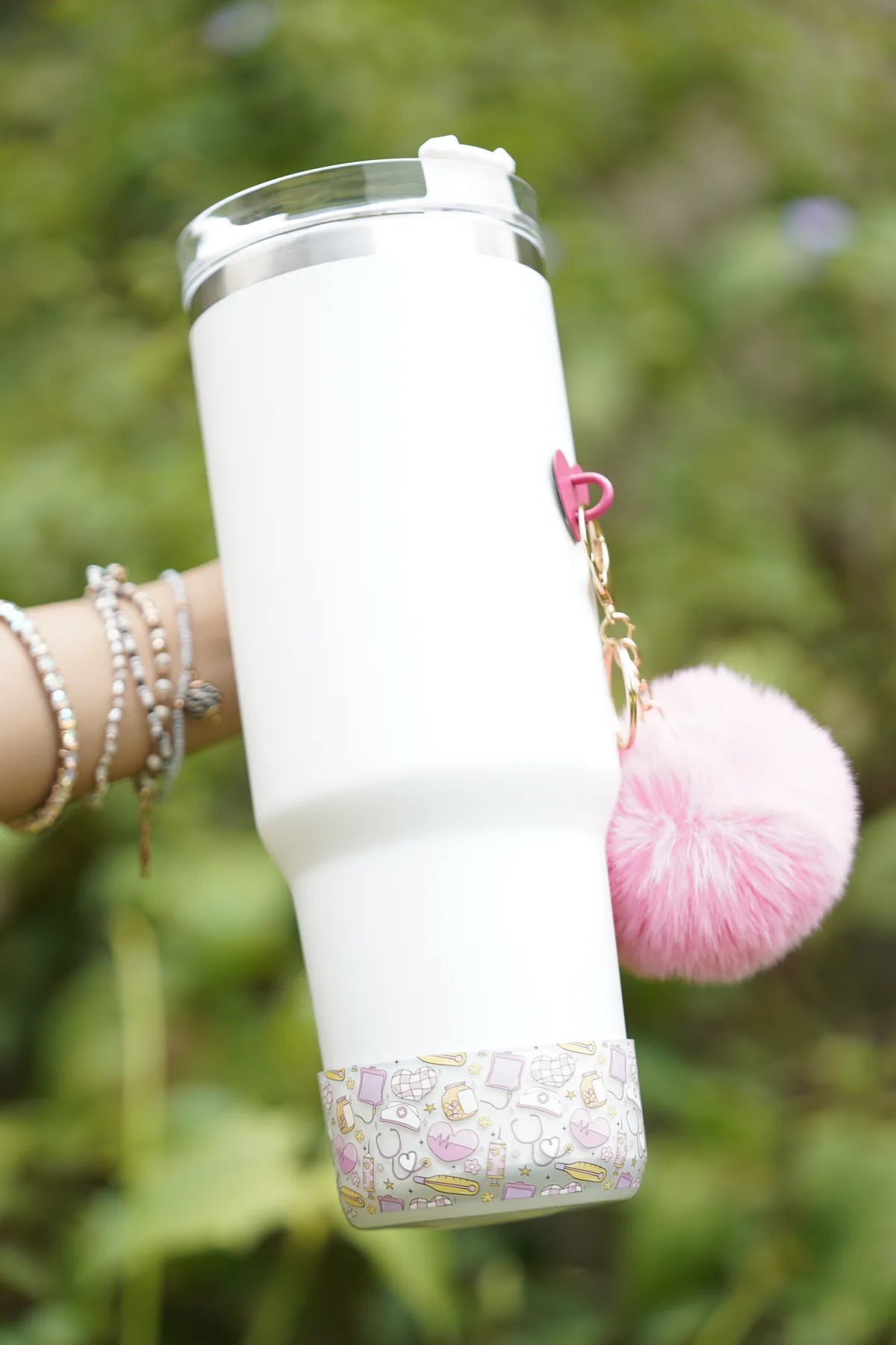 Nurse Chic water bottle boot fitted around the base of a white water bottle, featuring a pastel medical-themed pattern and a fluffy pink charm, shown outdoors.