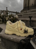 Close-up beige sneaker featuring black spider and ruby red glitter star charms with silver chain